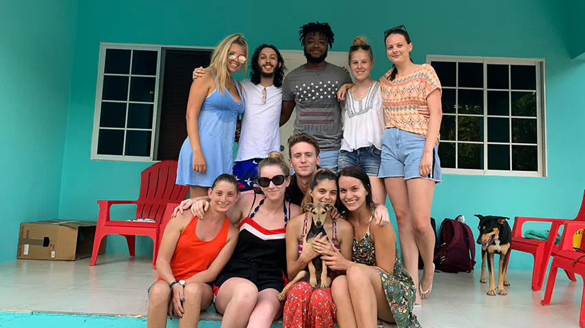 A group of ten diverse volunteers pose together outdoors in front of a turquoise wall. They showcase a friendly atmosphere, with some holding a small dog. Red chairs are visible in the background, highlighting a vibrant volunteer experience