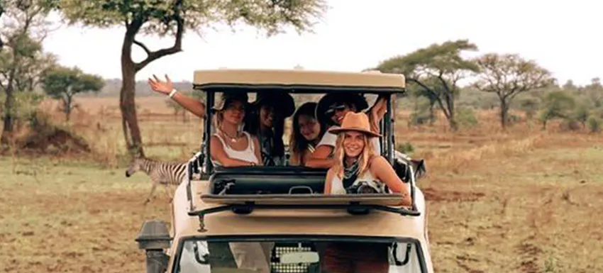Group of five volunteer travelers enjoying a safari adventure in a windowless safari vehicle. They are smiling, waving, and wearing hats in a scenic landscape with trees and grassland. A zebra is visible near the vehicle