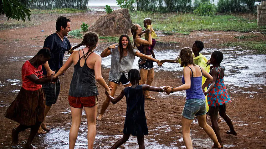 Five volunteer travelers and a group of local children joyfully dance in the rain, creating a lively atmosphere in a rural setting. The scene showcases cultural exchange and community bonding, highlighting the spirit of togetherness and fun