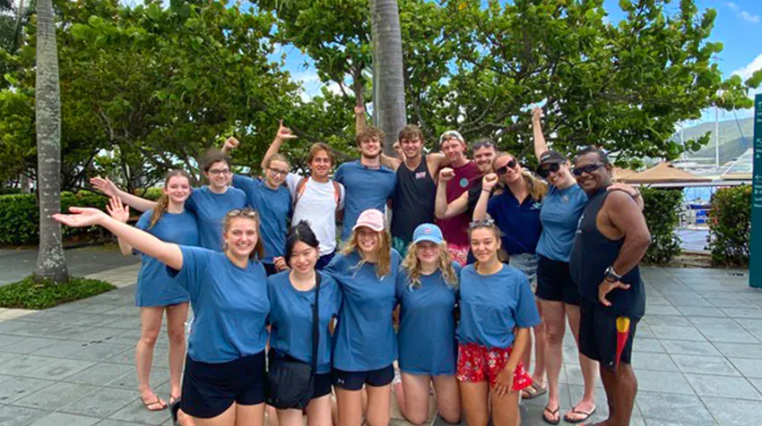 A large group of volunteer travelers in blue shirts pose for a photo outdoors with arms outstretched