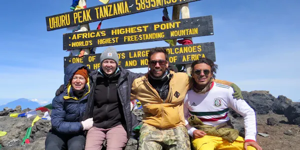 A group of four climbers who are international volunteers sit together, smiling, in front of the Mount Kilimanjaro summit sign at Uhuru Peak, Tanzania, which marks 5,895 meters (19,341 feet) above sea level.