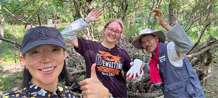 Two female volunteers smiling and posing together with a local team member in a forest in Soul, Korea. Wearing gloves and outdoor gear, showing enthusiasm for their conservation work.