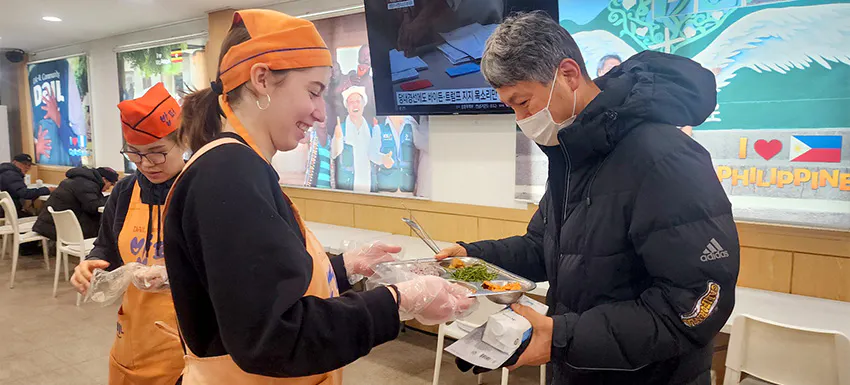 A volunteer wearing an orange apron and hat serves a warm meal to a community member in South Korea. Other volunteers are also engaged in meal distribution in the background.