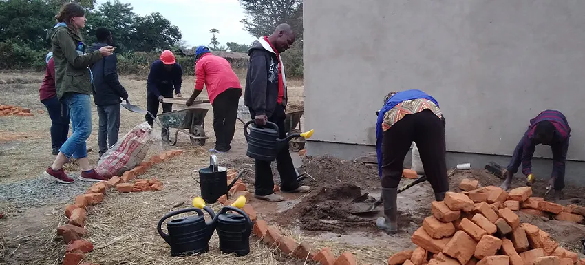 Volunteers and local community members work together on a construction and renovation project in Malawi, laying bricks, digging, and carrying materials to help build and improve infrastructure.