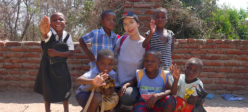 A volunteer poses with a group of smiling children in Malawi, all waving at the camera as they stand in front of a brick wall.