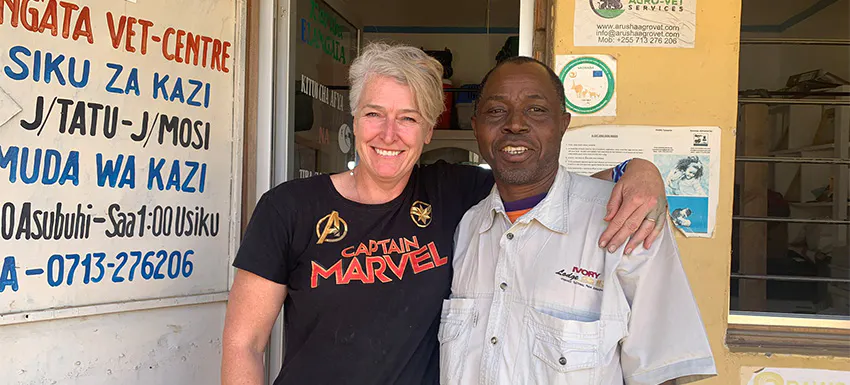 A smiling volunteer in a 'Captain Marvel' t-shirt stands arm-in-arm with a local veterinary worker outside a veterinary center, with a sign displaying work hours in Swahili.