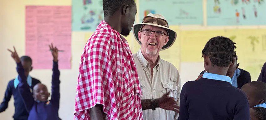 An older male volunteer wearing a hat and glasses smiles while engaging in conversation with a local teacher in a checkered cloth and schoolchildren in a brightly decorated classroom