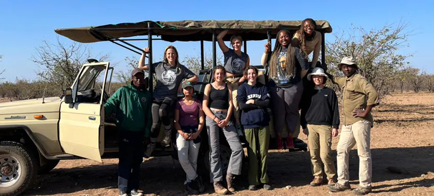 A group of volunteers and local guides pose together in front of an open safari vehicle in a dry, savannah-like landscape, dressed in outdoor gear and ready for an adventure.