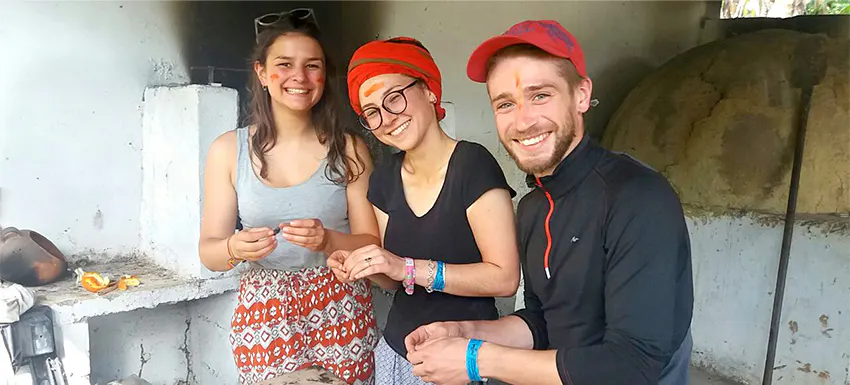 Three smiling volunteers, wearing casual clothing and headbands, stand together in a outdoor kitchen, preparing food and engaging in a cultural cooking experience.