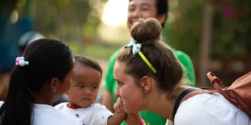 An IVHQ volunteer moment: a young woman with a bun and a blue bow leans in to talk to a curious toddler, while a local caregiver watches. Outdoors, on a sunny day, bracelets and a backpack highlight cross-cultural kids' outreach and community volunteering.