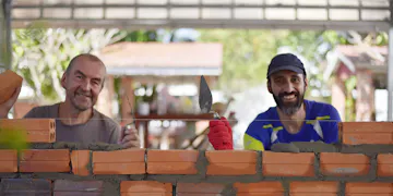Two volunteers build a brick wall outdoors. They smile at the camera, one with a trowel, the other wearing a cap and red glove. Bricks and fresh mortar form a wall in progress—teamwork on a community construction project.