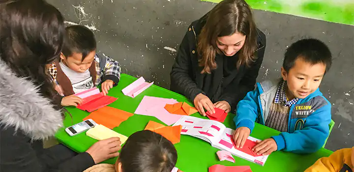 Two volunteers help young children fold colorful origami paper at a small green table, all focused on a step-by-step guide during a classroom activity in Guanxi, China.