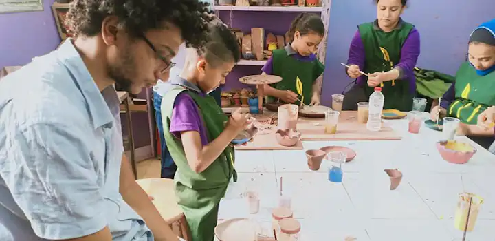 A childcare volunteer in Cairo, Egypt through the IVHQ childcare project and a group of children sit around a table carefully painting clay pottery pieces in a classroom filled with colorful materials and ceramic artworks.