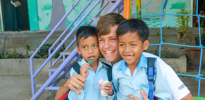 A young volunteer smiles with two local children in matching light blue school uniforms. They are outside a colorful childcare playground in Senggigi, Lombok - with one child holding a treat.