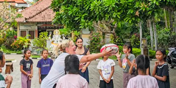 A sports education volunteer engages with local senggigi children in a playful rugby game in a vibrant outdoor setting, set against lush greenery and traditional architecture