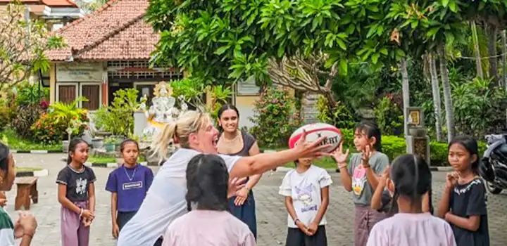 A sports education volunteer engages with local senggigi children in a playful rugby game in a vibrant outdoor setting, set against lush greenery and traditional architecture