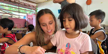 A volunteer teacher is pointing at a page in a notebook, helping the girl with what appears to be schoolwork or reading practice. The girl is attentively looking at the notebook.
