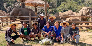 A group of international volunteers and local staff kneel and sit together, smiling for a photo in front of a wooden fence, with elephants and forested hills in the background at an elephant sanctuary in Chiang Mai, Thailand.